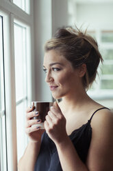 Porträt einer lächelnden blonden Frau mit einem Glas Kaffee, die durch ein Fenster schaut - NAF00067