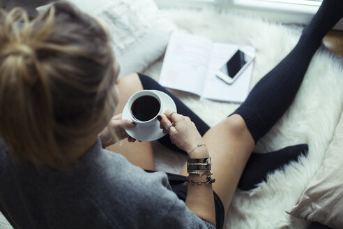 Young woman with cup of coffee relaxing on sheepskin at home - NAF00070