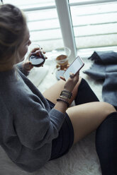 Junge Frau mit einer Tasse Kaffee, die sich zu Hause auf einem Schaffell ausruht und auf ihr Mobiltelefon schaut - NAF00073