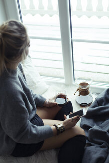 Young woman with cup of coffee and smartphone sitting on sheepskin at home looking through window - NAF00074