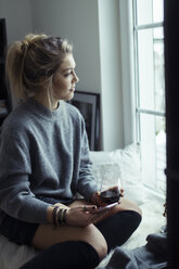 Young woman with cup of coffee and smartphone looking through window - NAF00075