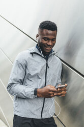 Athlete at building front with earbuds looking at cell phone - BOYF00675