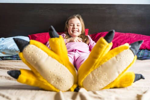 Smiling girl lying on bed wearing yellow fun slippers - XCF00150