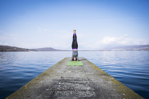 Frau übt Yoga und macht einen Kopfstand auf einem Pier an einem See, lizenzfreies Stockfoto