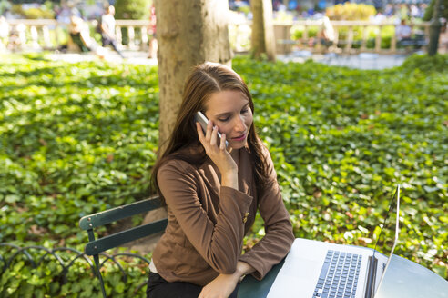 Junge Geschäftsfrau am Telefon sitzt am Tisch in einem Park mit Laptop - BOYF00696