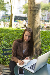 USA, New York, Manhattan, junge Geschäftsfrau mit Laptop sitzt an einem Tisch in einem Park und macht sich Notizen - BOYF00700