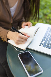 Junge Geschäftsfrau mit Laptop sitzt an einem Tisch in einem Park und macht sich Notizen, Teilansicht - BOYF00701