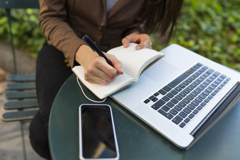 Junge Geschäftsfrau mit Laptop sitzt an einem Tisch in einem Park und macht sich Notizen, Teilansicht, lizenzfreies Stockfoto