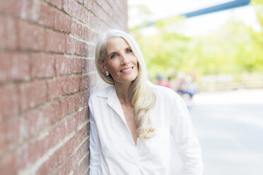 Portrait of smiling mature woman wearing white shirt blouse leaning against brick wall - GIOF02292