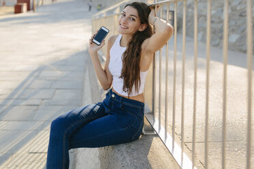 Portrait of smiling young woman with smartphone sitting on a wall - GIOF02325