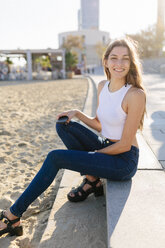 Spain, Barcelona, portrait of smiling young woman sitting on beach promenade at sunset - GIOF02329