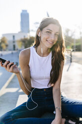 Spain, Barcelona, portrait of smiling young woman relaxing at sunset listening music with earphones - GIOF02337