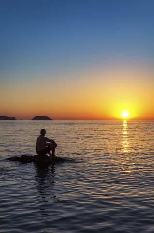 Spanien, Menorca, Playa de Cavalleria, Mann auf Felsen im Meer bei Sonnenuntergang, lizenzfreies Stockfoto