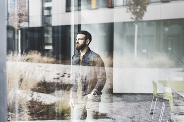 Young businessman standing at window pane, wearing smart watch - UUF10156