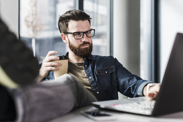 Young casual businessman sitting in office with feet up and a cup of coffee - UUF10163