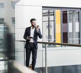 Businessman standing in office building, using smart phone and digital tablet - UUF10184