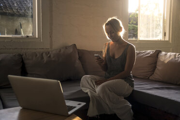 Blonde Frau sitzt auf der Couch im Gegenlicht und schaut auf ihr Handy - KNTF00813