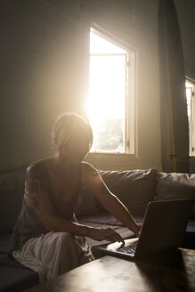 Blonde Frau sitzt auf der Couch bei Gegenlicht und benutzt einen Laptop - KNTF00818