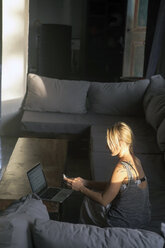 Blonde Frau sitzt auf der Couch bei Sonnenlicht und benutzt ein Handy - KNTF00819