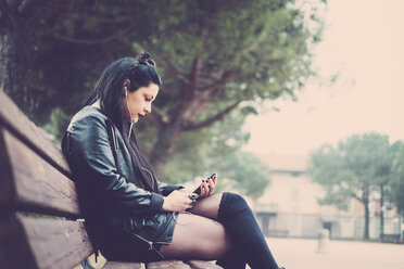 Dark-haired young woman sitting on bench listening music with earphones and smartphone - SIPF01484