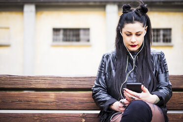 Dunkelhaarige junge Frau sitzt auf einer Bank und hört mit Kopfhörern und Smartphone Musik - SIPF01485