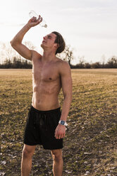 Barechested athlete in rural landscape pouring water over his face - UUF10218