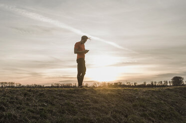 Sportler schaut auf Handy in ländlicher Landschaft bei Sonnenuntergang - UUF10232