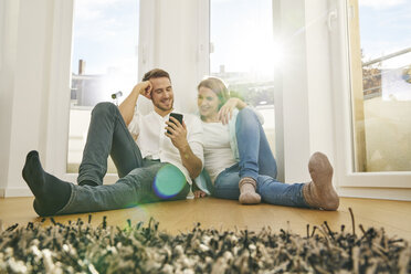 Lächelndes Paar sitzt auf dem Boden und schaut auf sein Handy - FMKF03603
