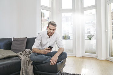 Mann sitzt auf der Couch und schaut auf sein Handy - FMKF03624