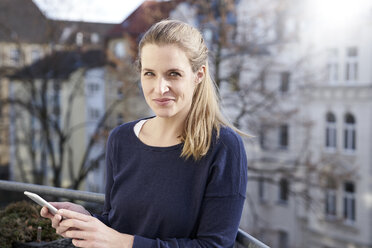 Porträt einer lächelnden Frau mit Mobiltelefon auf einem Balkon - FMKF03637