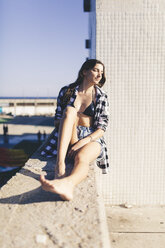 Young pretty woman wearing beach wear sitting on wall - GIOF02351