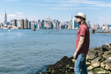 USA, New York City, Mann mit Hut am Wasser mit der Skyline von Manhattan im Hintergrund - GIOF02395