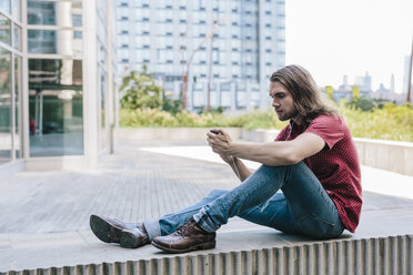 Man sitting on a wall with earbuds using cell phone - GIOF02405