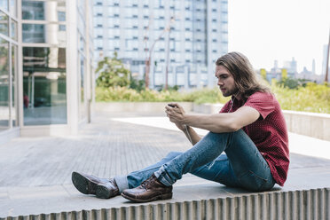 Mann sitzt mit Ohrstöpseln an einer Wand und benutzt ein Mobiltelefon - GIOF02405