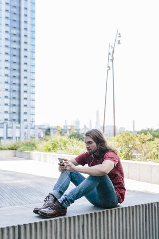 Mann sitzt mit Ohrstöpseln an einer Wand und benutzt ein Mobiltelefon, lizenzfreies Stockfoto
