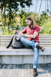 Man sitting on a bench with earbuds and backpack - GIOF02409