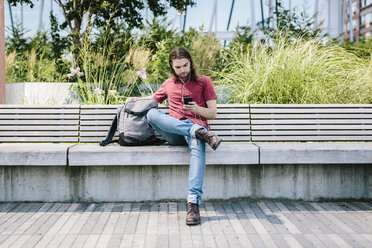 Man sitting on a bench with earbuds using cell phone - GIOF02410