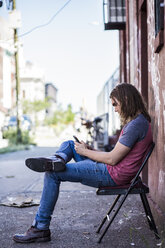 USA, New York City, Mann sitzt auf einem Stuhl und benutzt ein Mobiltelefon in Williamsburg, Brooklyn - GIOF02428