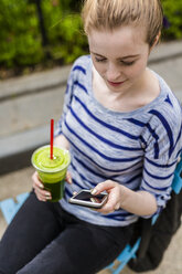 Junge Frau trinkt einen Smoothie und überprüft ihr Handy im Freien - GIOF02447