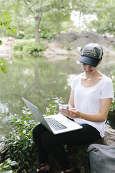 Junge Frau sitzt am Seeufer im Park und benutzt Handy und Laptop - BOYF00746