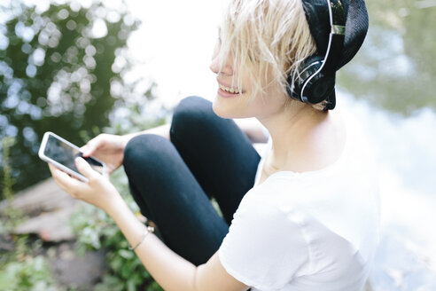 Junge Frau sitzt am Seeufer im Park, trägt Kopfhörer und hält ein Handy in der Hand - BOYF00750
