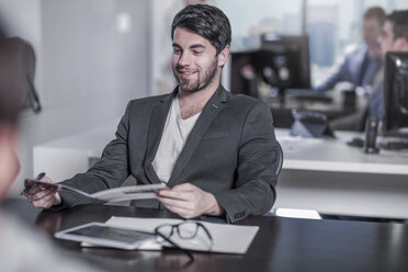 Smiling man looking at booklet at desk in city office - ZEF13197