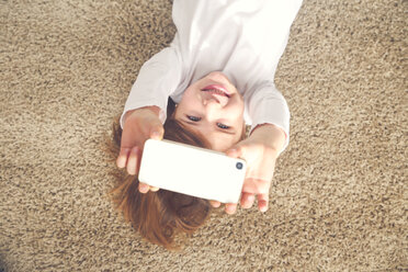 Porträt eines grinsenden kleinen Mädchens, das auf dem Teppich liegt und ein Selfie mit seinem Smartphone macht - RTBF00756