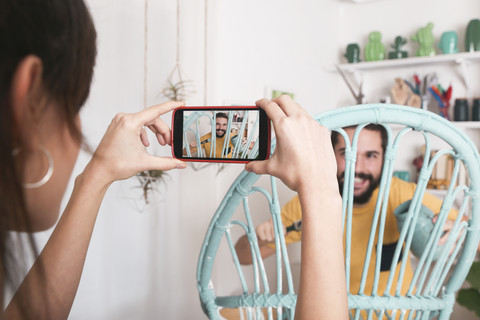 Junge Frau, die einen bärtigen Mann fotografiert, der zu Hause einen Korbsessel bemalt, lizenzfreies Stockfoto