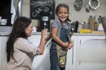 Mother tying apron for daughter in kitchen - ZEF13205