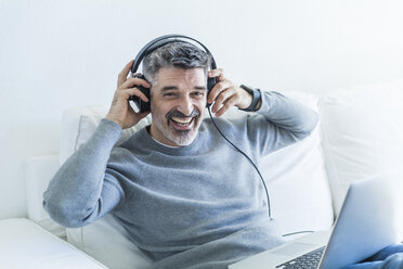 Portrait of happy mature man at home using laptop and wearing headphones - TCF05337