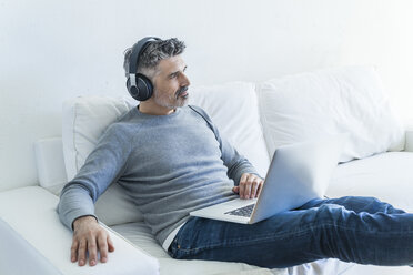 Mature man at home using laptop and wearing headphones - TCF05338