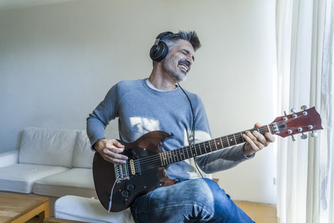 Älterer Mann spielt zu Hause E-Gitarre und trägt Kopfhörer, lizenzfreies Stockfoto