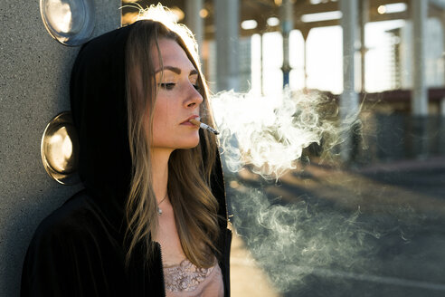 Porträt einer jungen Frau mit Kapuzenjacke, die bei Gegenlicht eine Zigarette raucht - KKAF00555