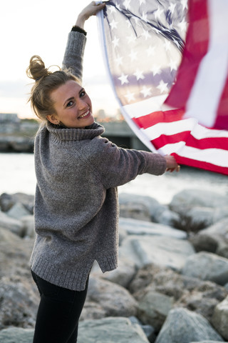 Porträt einer lächelnden jungen Frau mit wehender US-amerikanischer Flagge, lizenzfreies Stockfoto